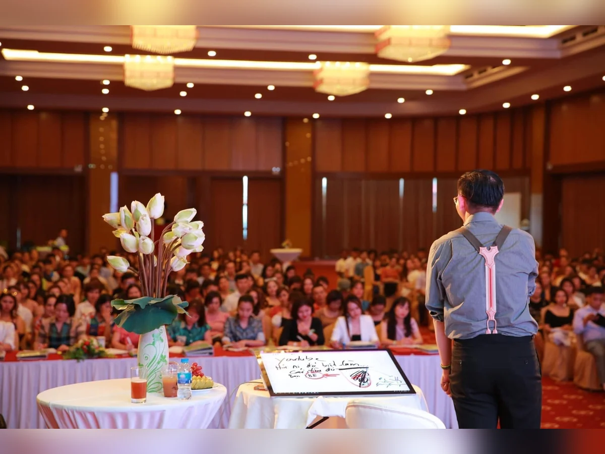 Diễn giả đứng trước bàn trình bày, hướng về hội trường đông người đang theo dõi.
