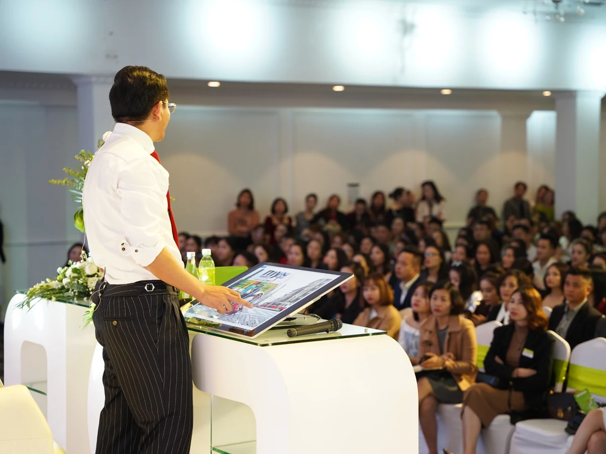 Diễn giả áo trắng đứng bên màn hình nghiêng trên bục, hướng về người tham dự trong hội trường.