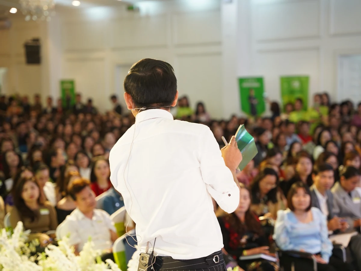 Diễn giả mặc áo trắng, đeo micro và cầm tài liệu khi hướng về đông người tham dự trong hội trường.
