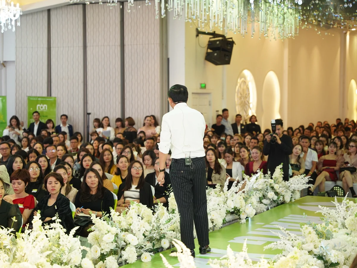 Diễn giả áo trắng đứng quay lưng về máy ảnh trước đông người tham dự, cạnh người đang chụp ảnh và hàng hoa trắng.