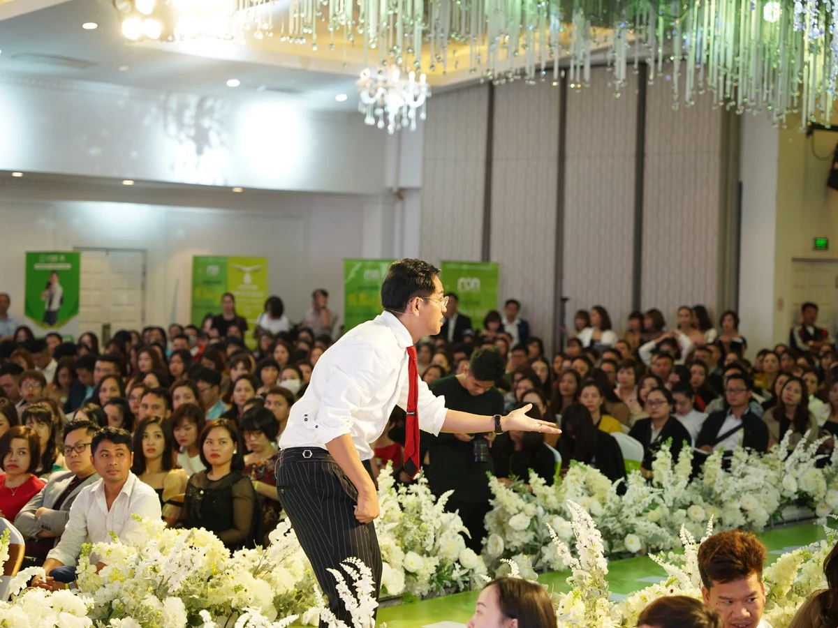 Diễn giả áo trắng và cà vạt đỏ đưa tay tương tác với người tham dự dọc lối sân khấu có hoa trắng.