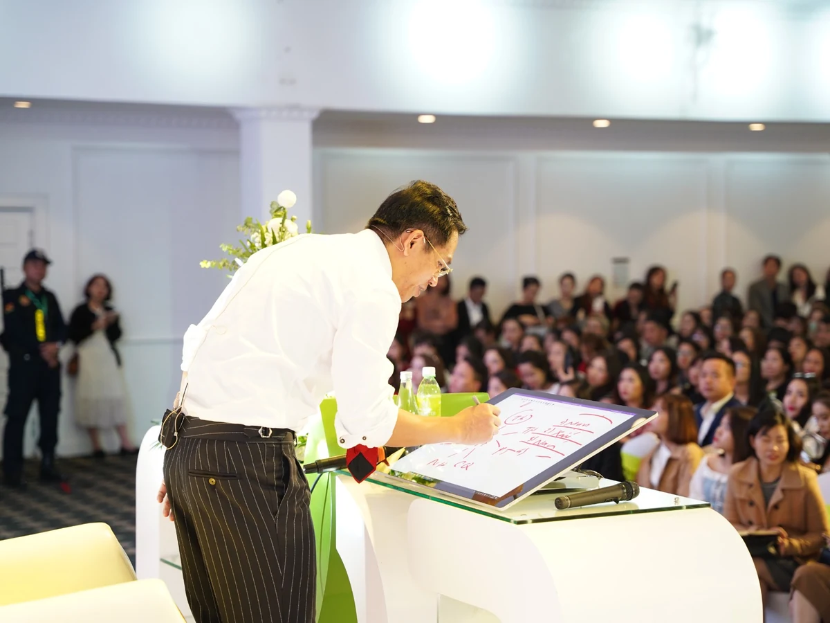 Diễn giả áo trắng viết bằng bút trên màn hình nghiêng đặt trên bục, trước người tham dự.