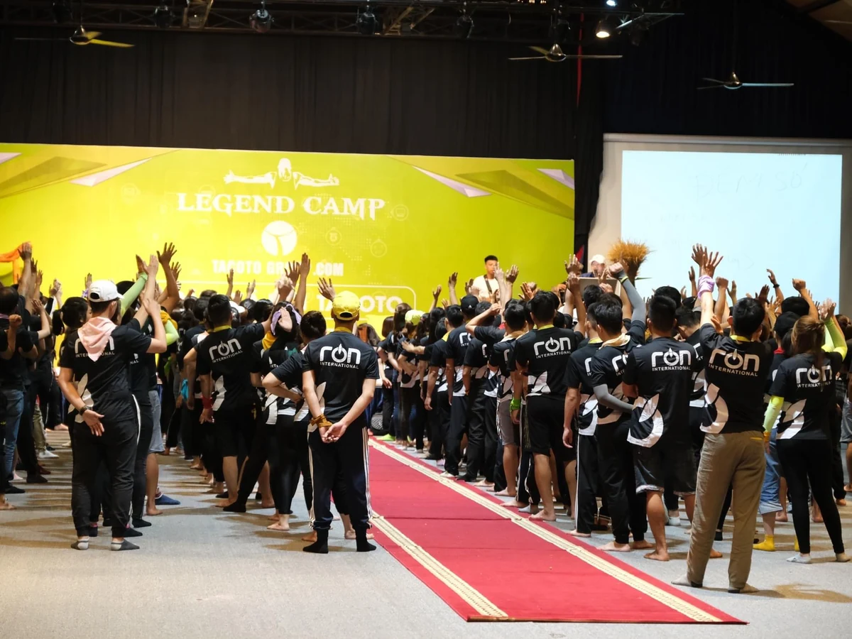 Nhóm người mặc áo đen có logo RON INTERNATIONAL giơ tay trước sân khấu LEGEND CAMP.