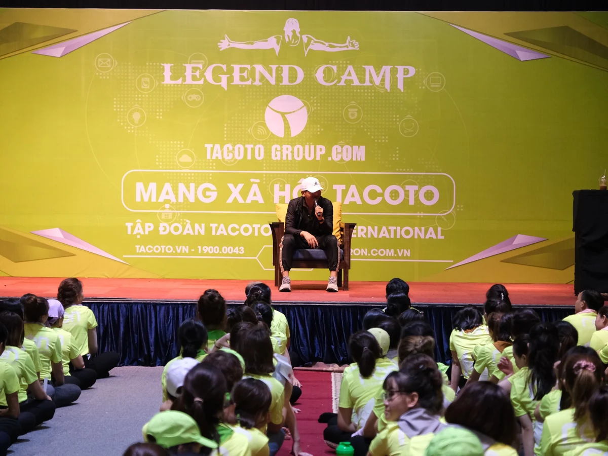 Người cầm micro ngồi trên ghế ở sân khấu LEGEND CAMP, phía trước là nhiều người tham dự mặc áo xanh vàng.