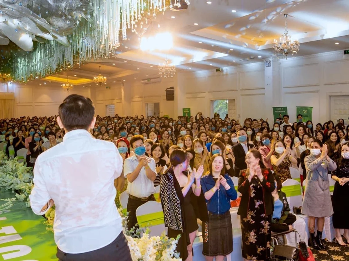 Người tham dự đứng vỗ tay hướng về diễn giả áo trắng trong hội trường trang trí hoa trắng.