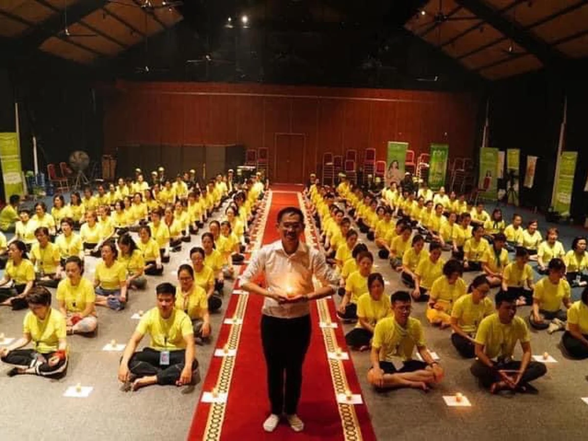 Nhóm người mặc áo xanh vàng ngồi xếp bằng thành hàng, một người đứng giữa lối thảm đỏ cầm vật phát sáng.