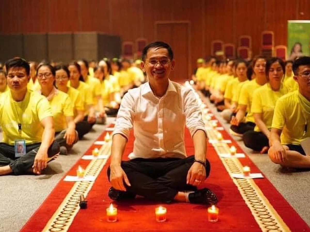 Một người áo trắng ngồi xếp bằng trên thảm đỏ phía trước hàng nến, cùng nhóm người áo xanh vàng ngồi phía sau.