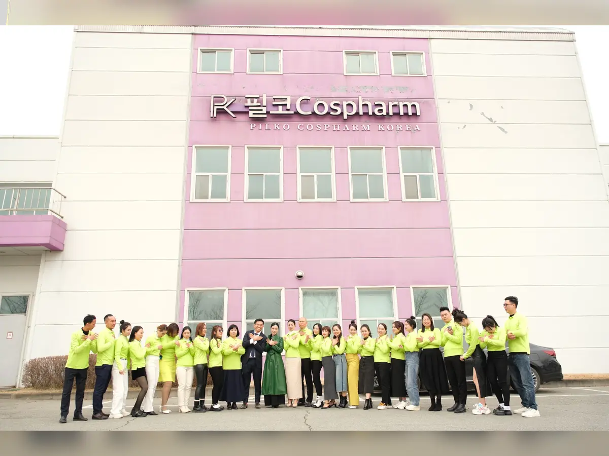 Đoàn người mặc áo xanh vàng chụp ảnh trước tòa nhà có biển PILKO COSPHARM KOREA.