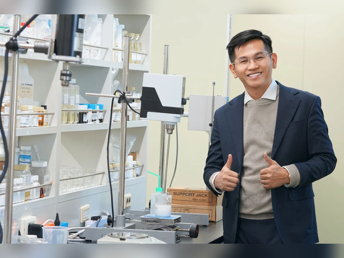 Người đàn ông mặc vest đứng cạnh thiết bị thí nghiệm và giơ hai ngón tay cái.