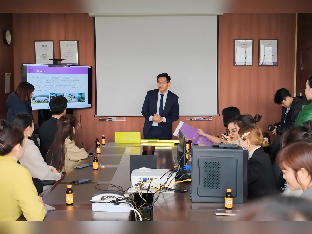 Người mặc vest chia sẻ trước nhóm đang ngồi quanh bàn họp có màn hình và máy chiếu.