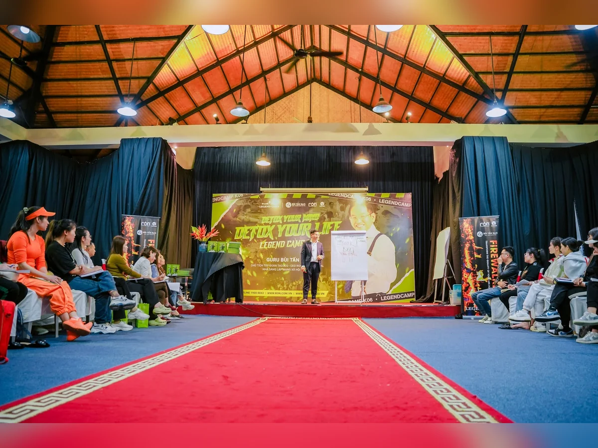 Diễn giả đứng cạnh bảng giấy trên sân khấu LEGEND CAMP, người tham dự ngồi hai bên lối thảm đỏ.