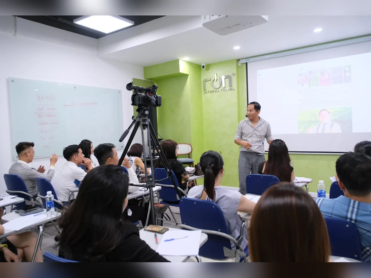 Diễn giả chia sẻ trước người tham dự trong phòng có logo RON INTERNATIONAL, màn chiếu và máy quay.