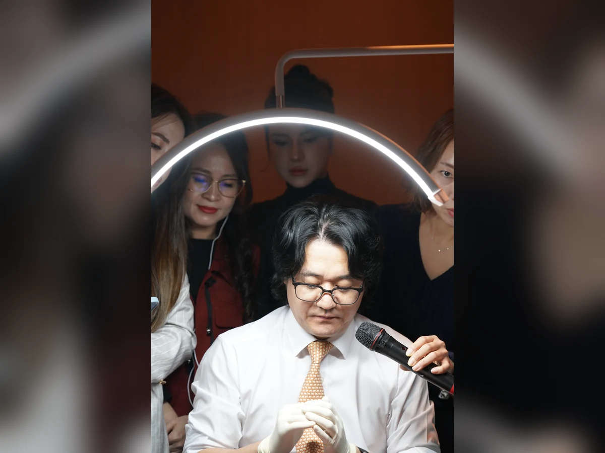 Người đàn ông đeo kính và găng tay ngồi dưới đèn vòng, bên cạnh có người đưa micro.