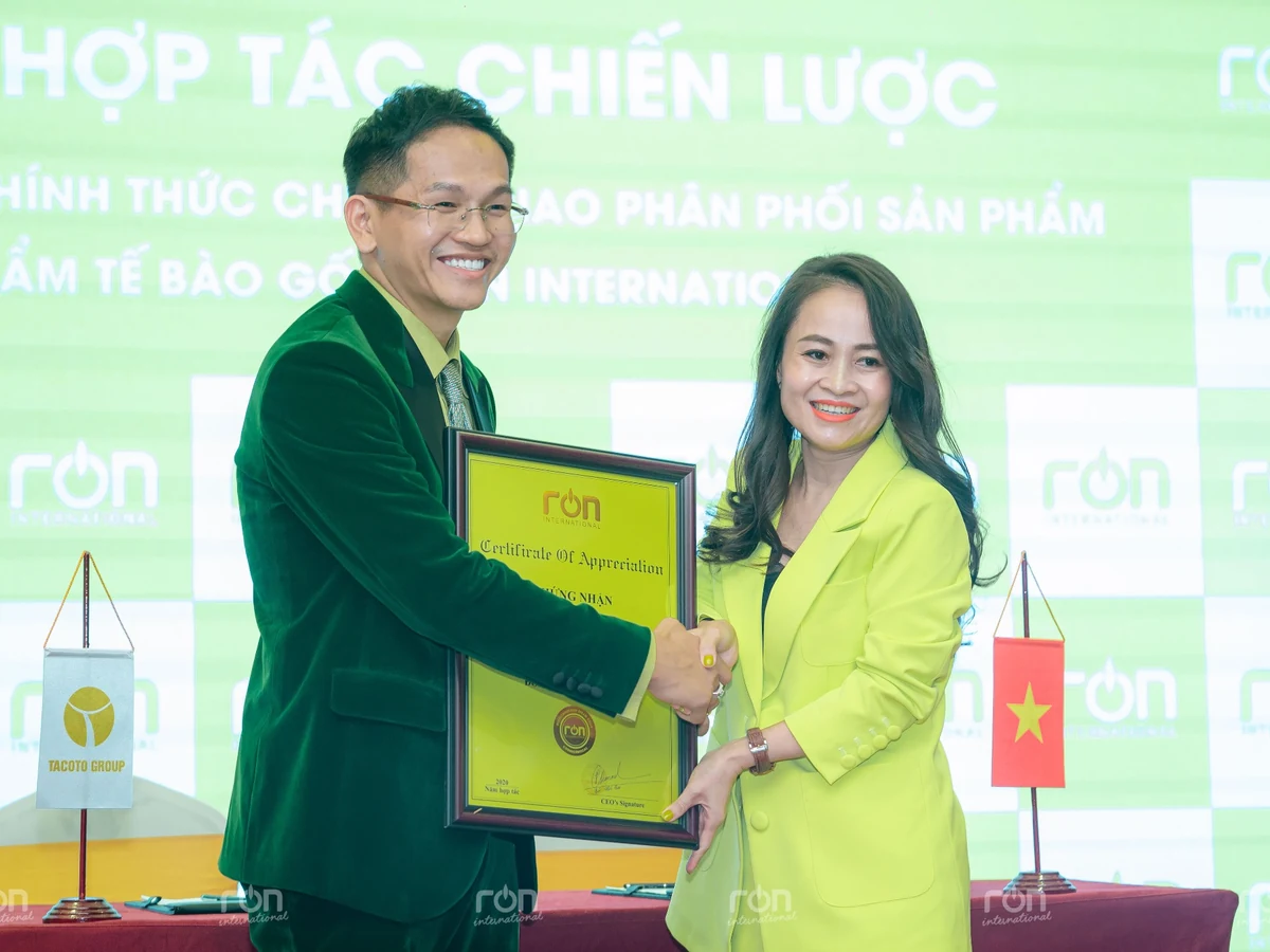 Hai người bắt tay và cùng cầm khung Certificate of Appreciation trước phông nền RON INTERNATIONAL.