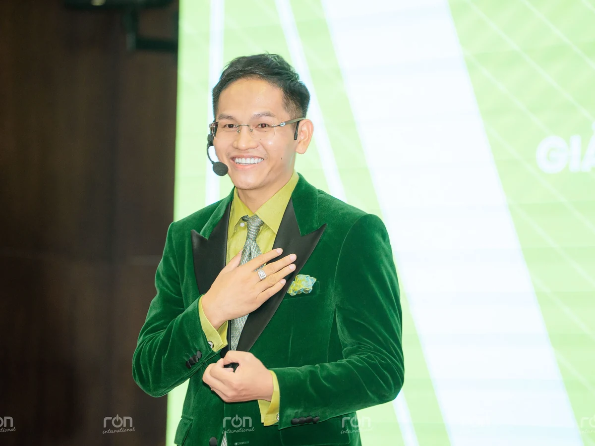 Diễn giả đeo micro, mặc vest xanh và mỉm cười trước phông nền xanh nhạt.
