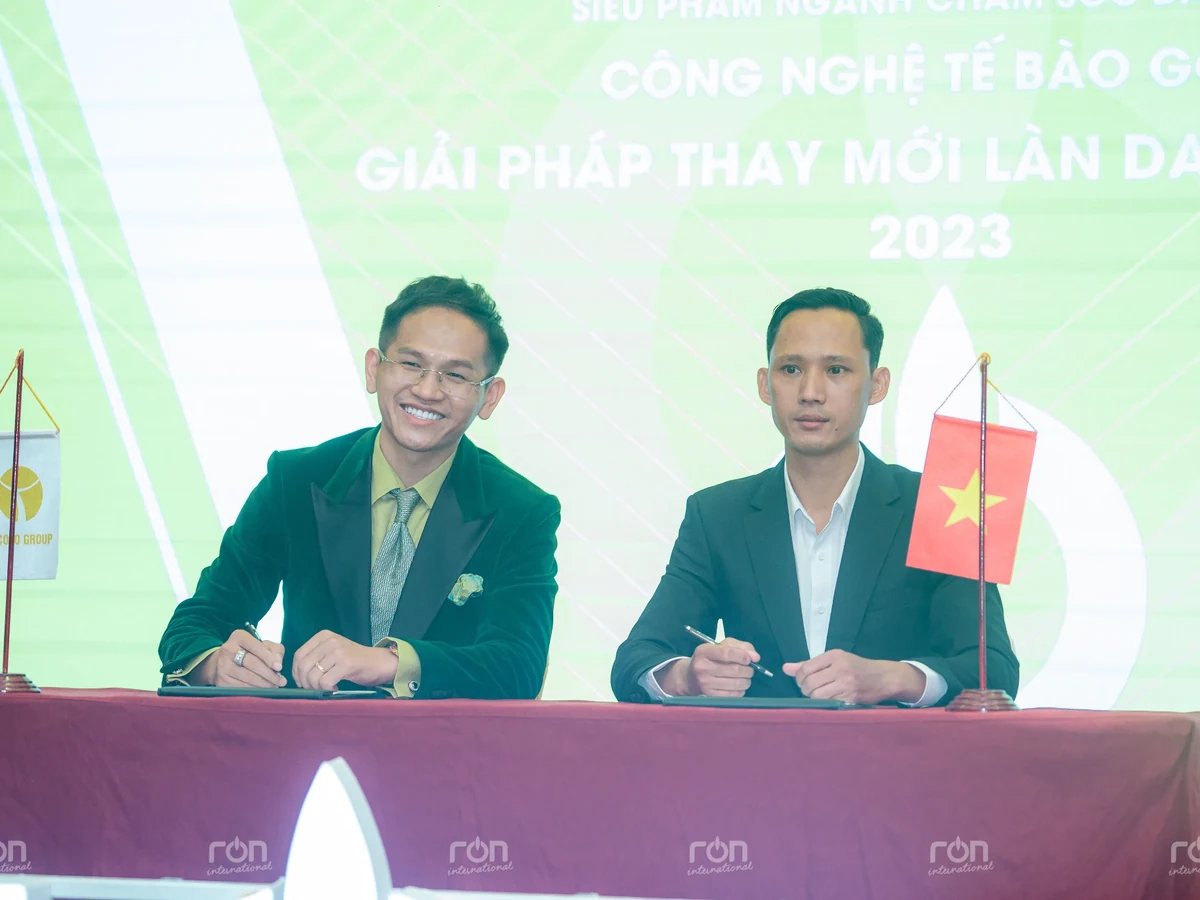 Hai người ngồi tại bàn cầm bút, trước phông nền xanh có năm 2023 và các cờ bàn.