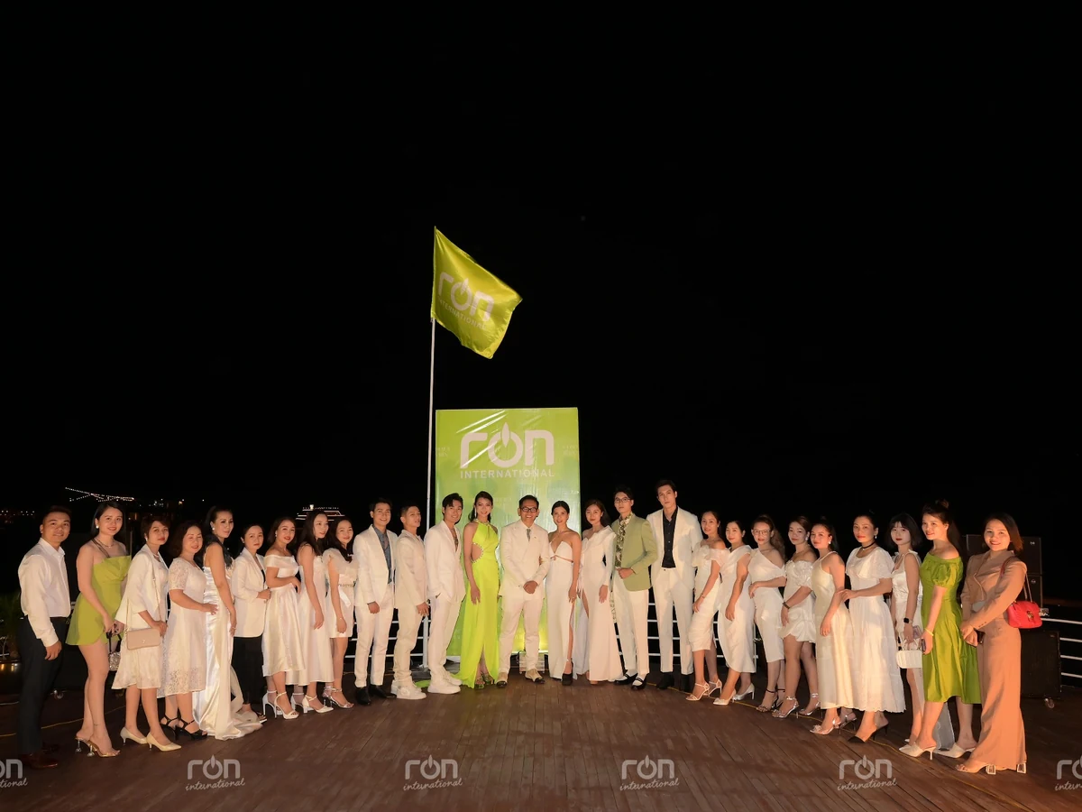 Đoàn người xếp hàng chụp ảnh buổi tối dưới cờ và bảng RON INTERNATIONAL, phần lớn mặc trang phục trắng.