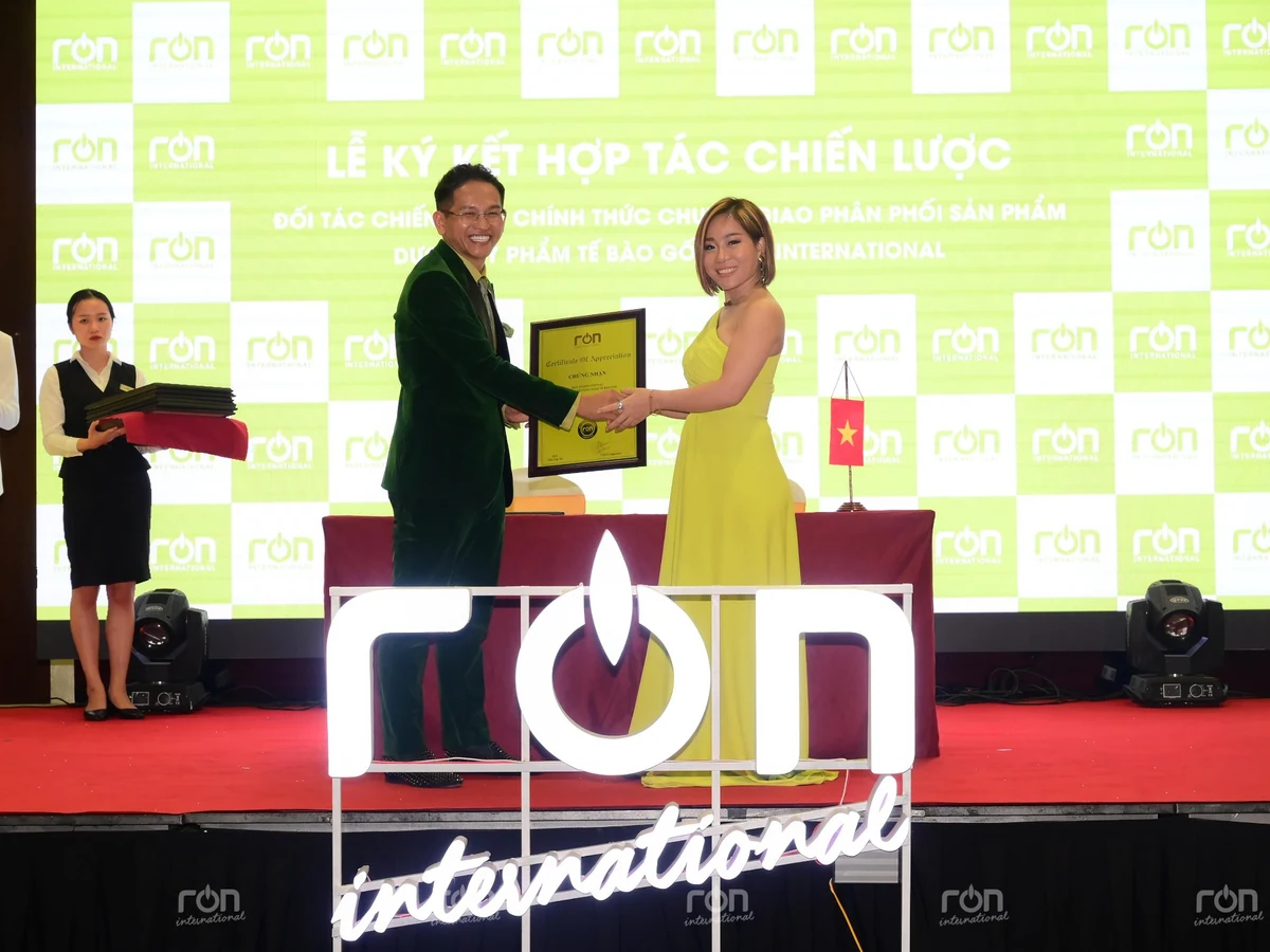 Hai người bắt tay và cùng cầm khung Certificate of Appreciation trên sân khấu có nhận diện RON INTERNATIONAL.