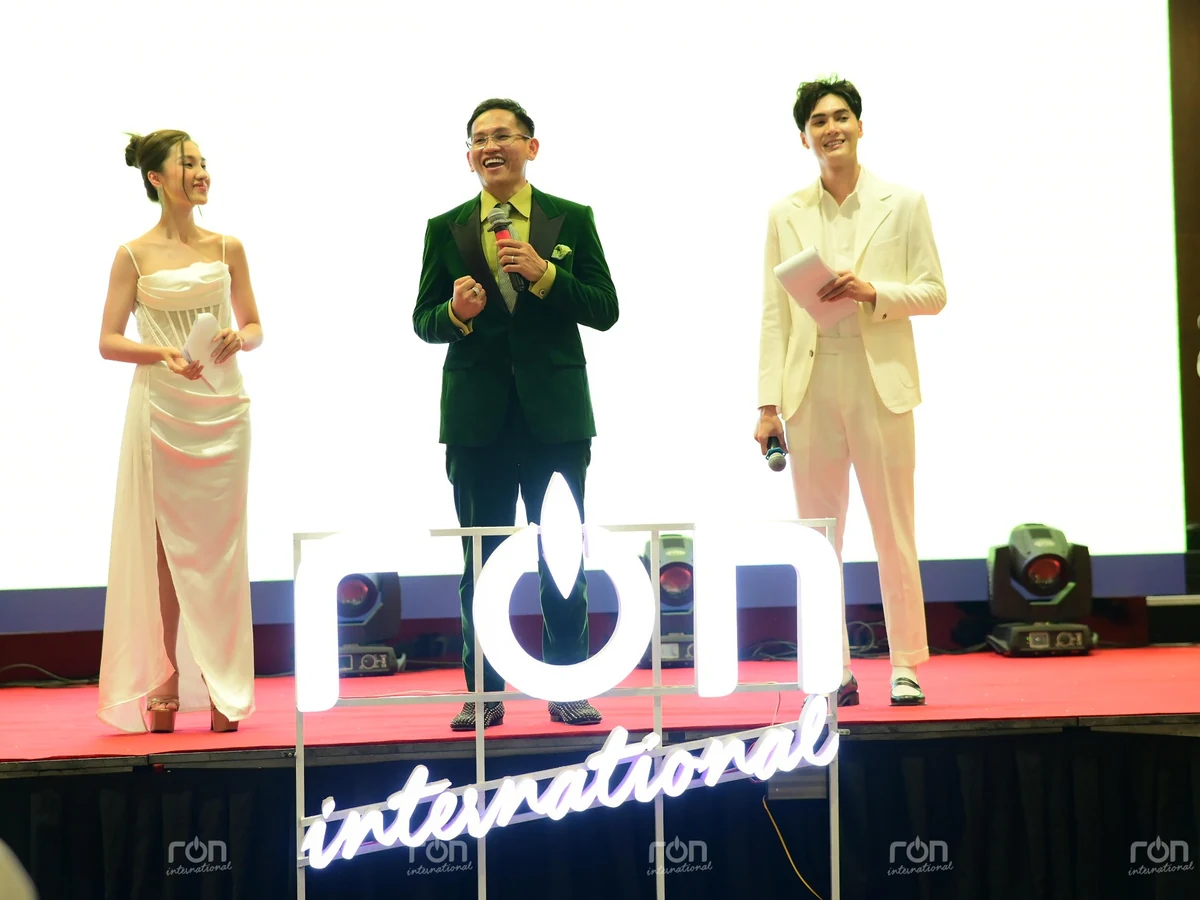 Ba người đứng trên sân khấu RON INTERNATIONAL; người giữa cầm micro, hai người hai bên cầm tập giấy.